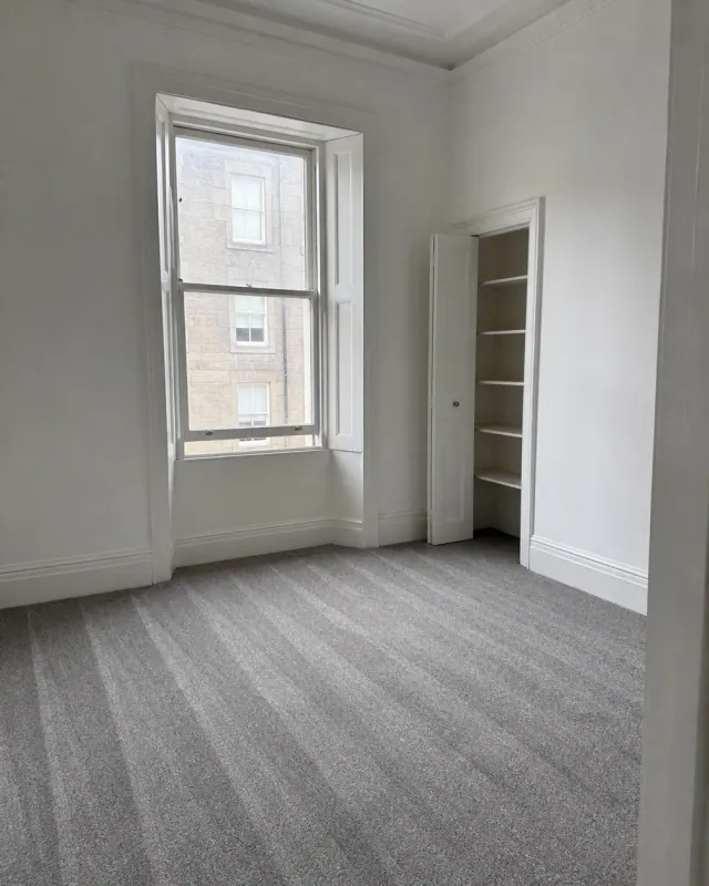 An empty bedroom with window shutters, an open press cupboard and fresh hoover lines in the carpet