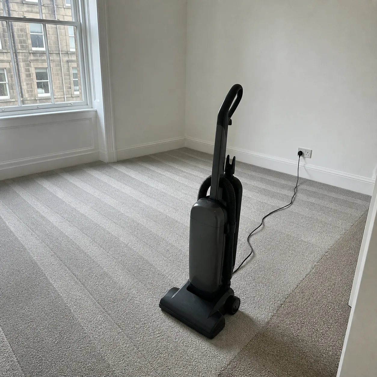 An upright vacuum standing in an empty bedroom with fresh hoover lines in the carpet